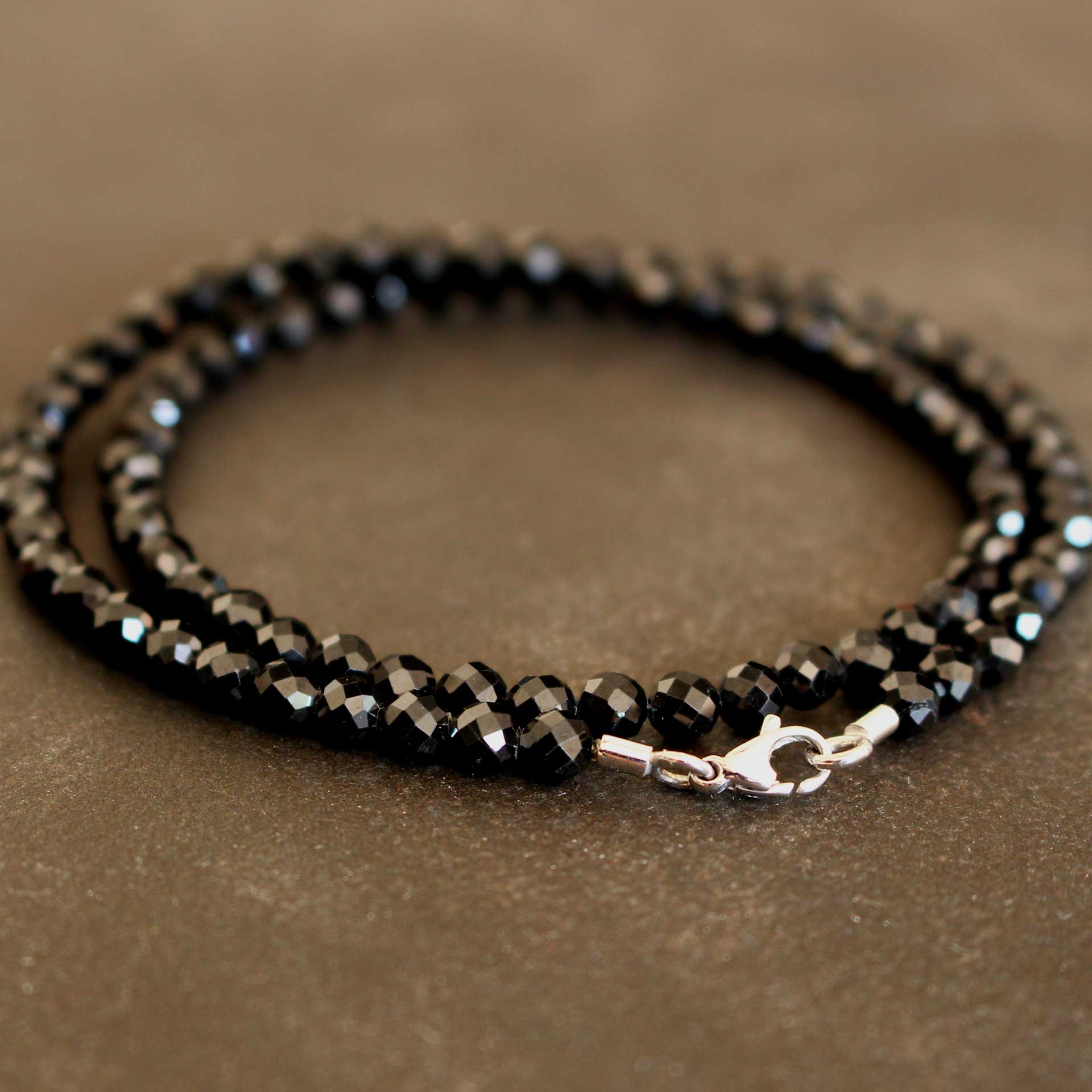 Nahaufnahme eines zweifach gewickelten Armbands aus facettierten schwarzen Spinell-Rondellen mit glänzendem Karabinerverschluss und Endtüllen aus 750er Weißgold auf braun-grauem Untergrund.