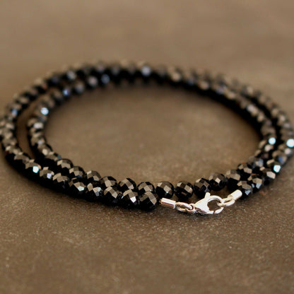 Nahaufnahme eines zweifach gewickelten Armbands aus facettierten schwarzen Spinell-Rondellen mit glänzendem Karabinerverschluss und Endtüllen aus 750er Weißgold auf braun-grauem Untergrund.