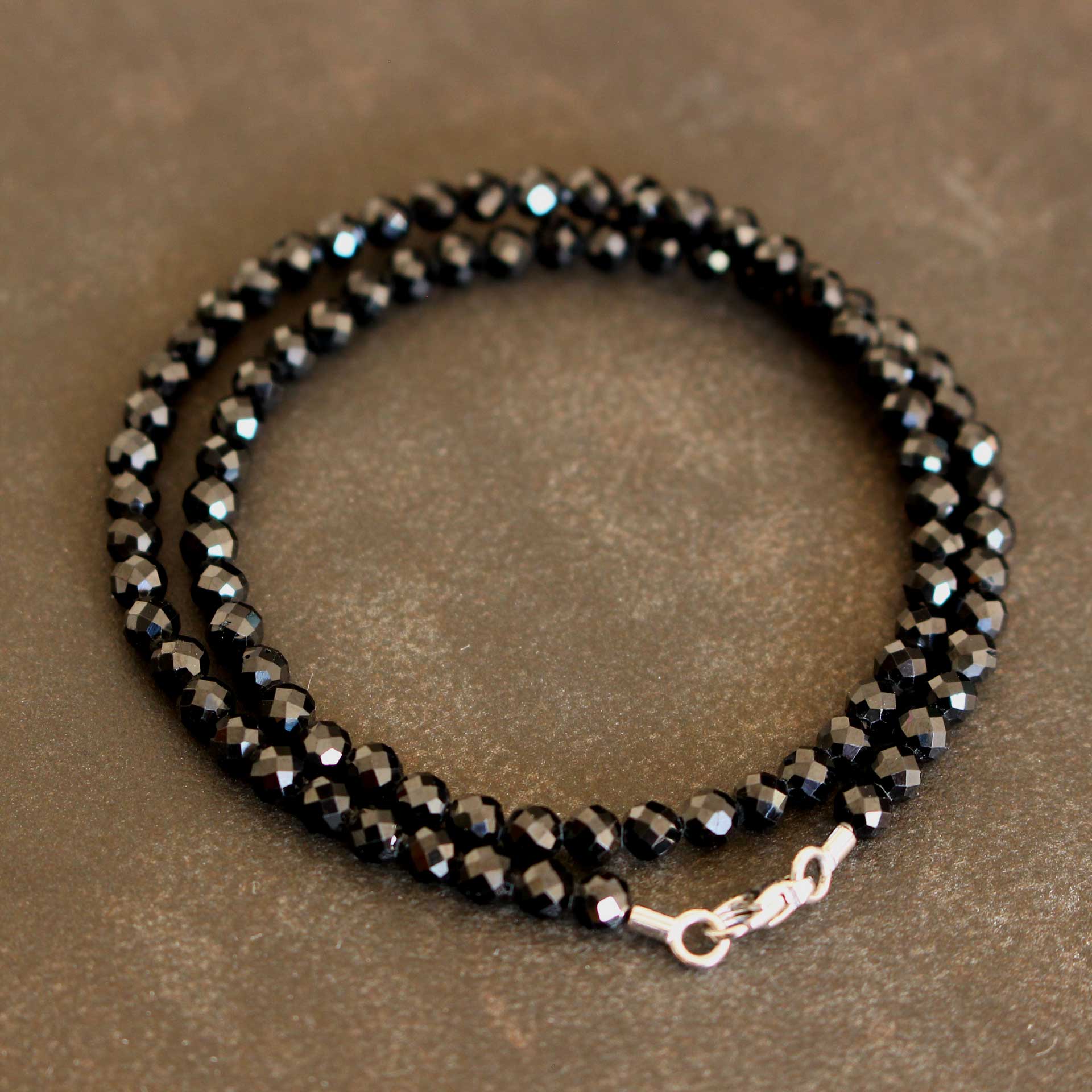 Nahaufnahme eines zweifach gewickelten Armbands aus facettierten schwarzen Spinell-Rondellen mit glänzendem Karabinerverschluss und Endtüllen aus 750er Weißgold auf braun-grauem Untergrund.